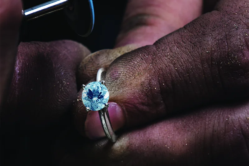 A blue gemstone ring being held under a jeweler's bench light